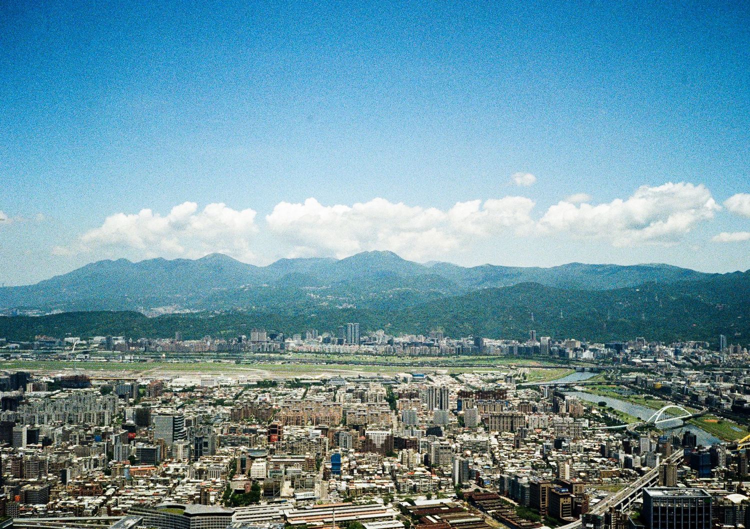 Panoramic cityscape view of an urban area surrounded by mountains under clear blue skies.