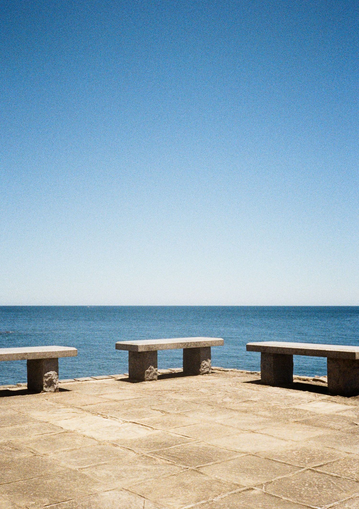 Concrete benches line a waterfront promenade overlooking the blue ocean horizon on a clear sunny day.