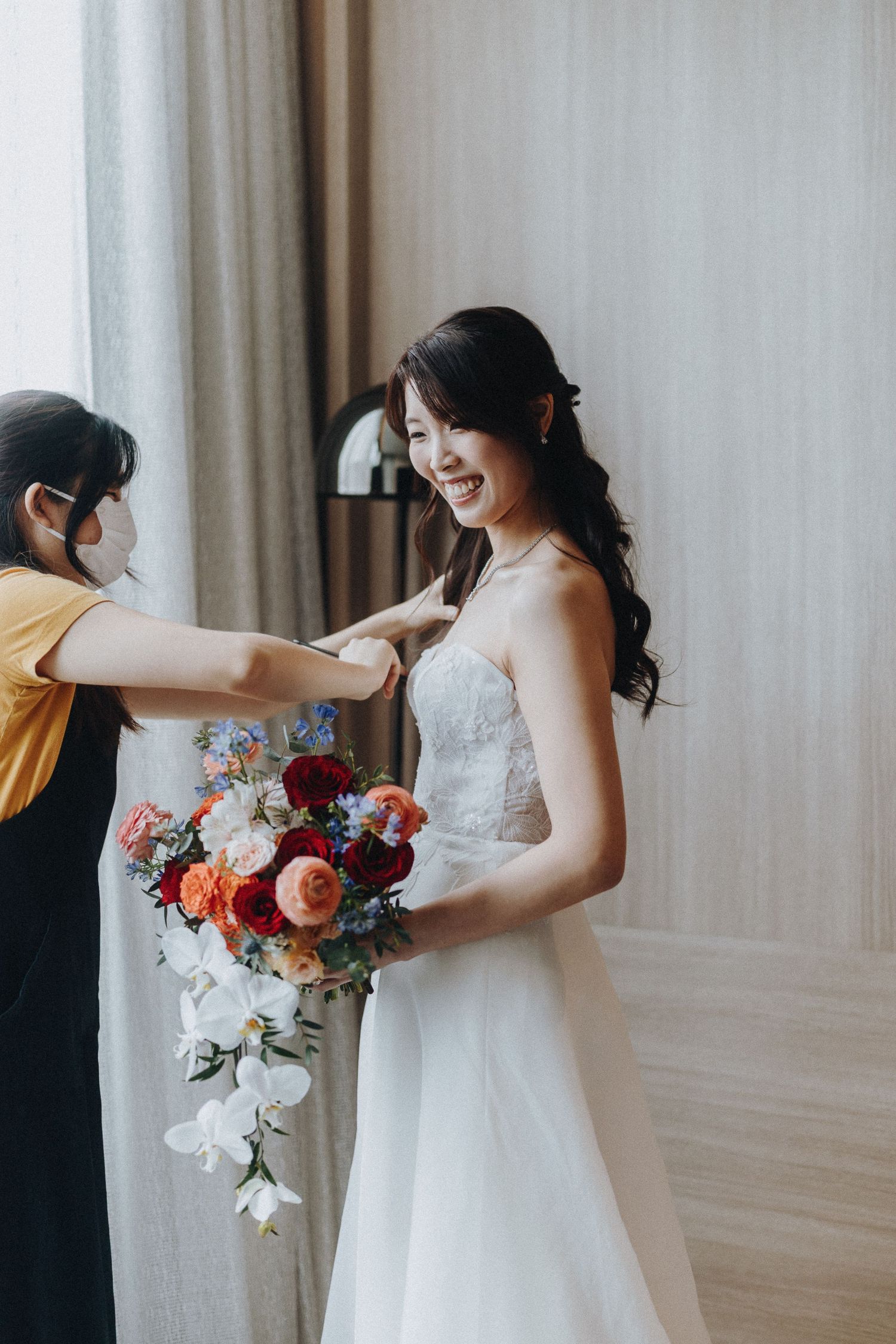 A bride in a white wedding dress holds a vibrant red and pink bouquet against a light-colored backdrop.
