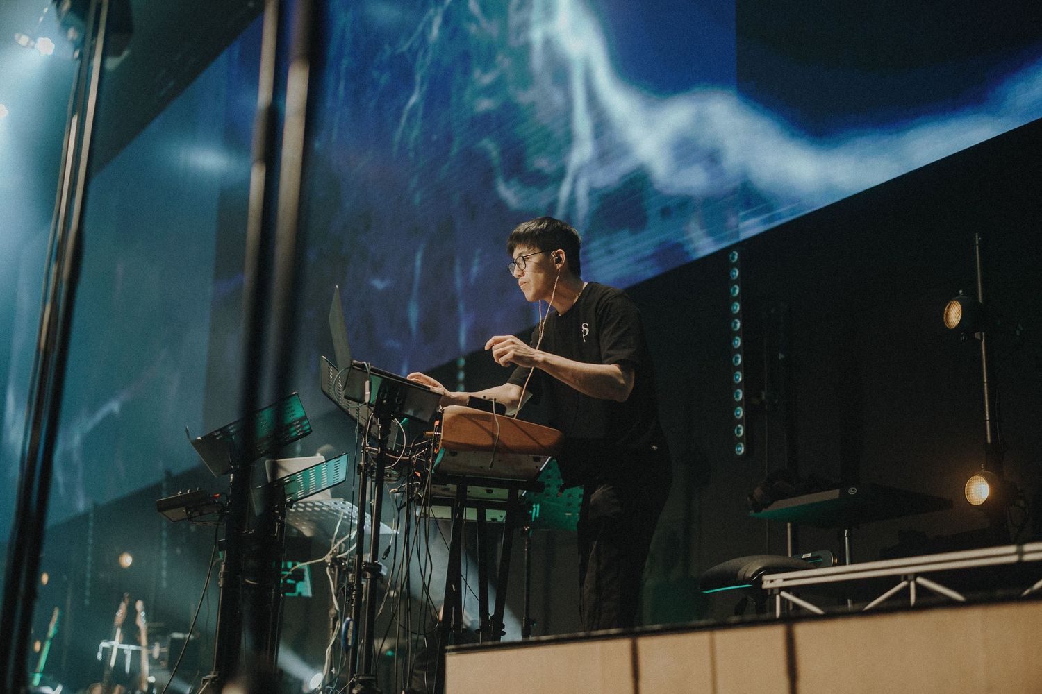 Drummer performs energetically on an elevated platform with dramatic blue lighting and stage effects in the background.