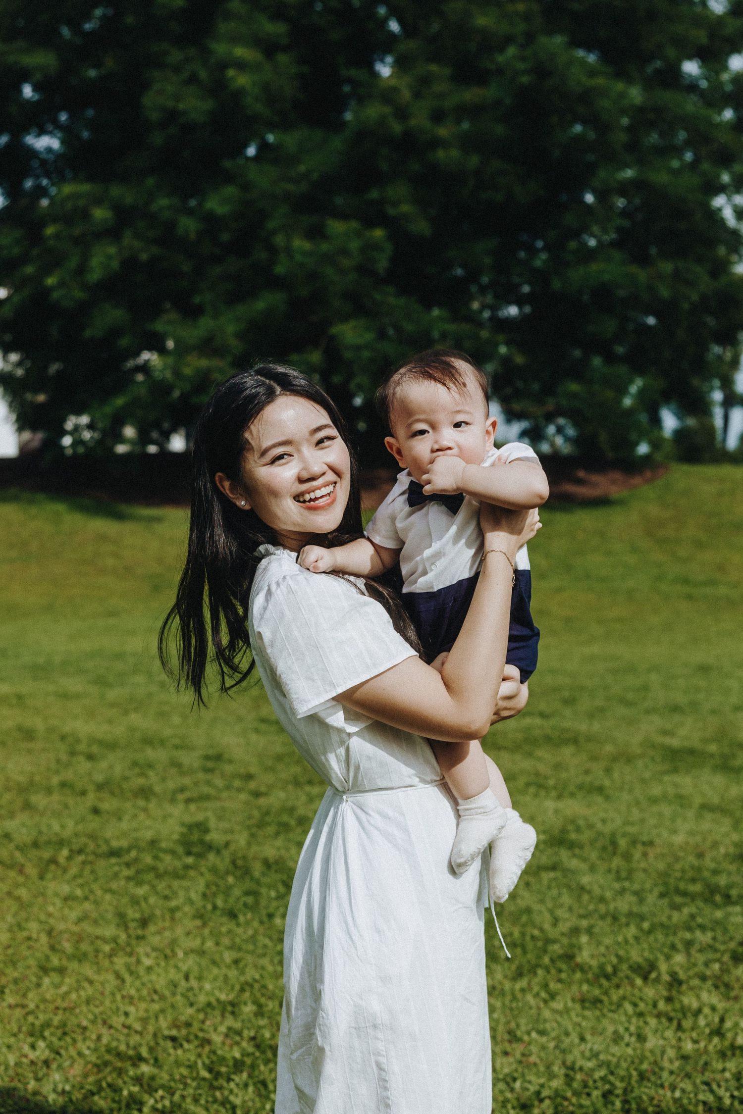 A tender outdoor moment shows a parent in a white dress holding and embracing a small child on a grassy hill.