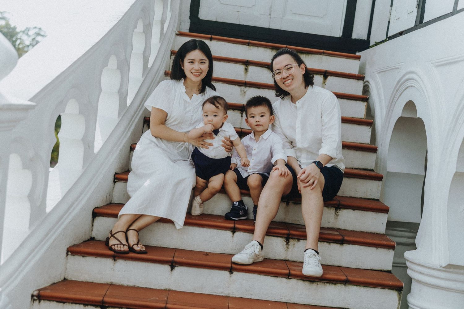 A family wearing coordinated white outfits sits together on a white staircase in elegant minimalist setting.