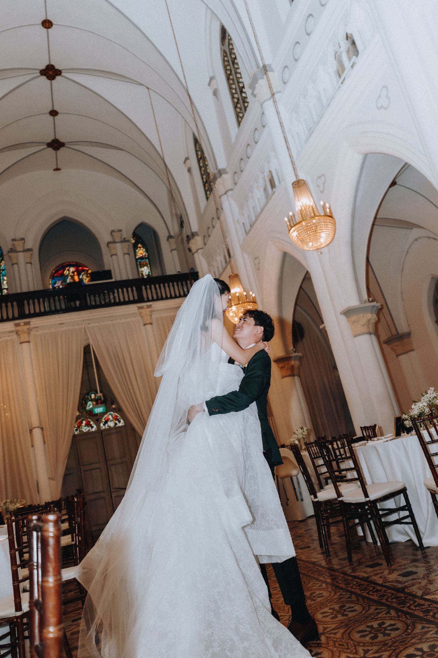 Bride and groom share a romantic embrace inside an elegant white church with arched ceilings and stained glass windows.