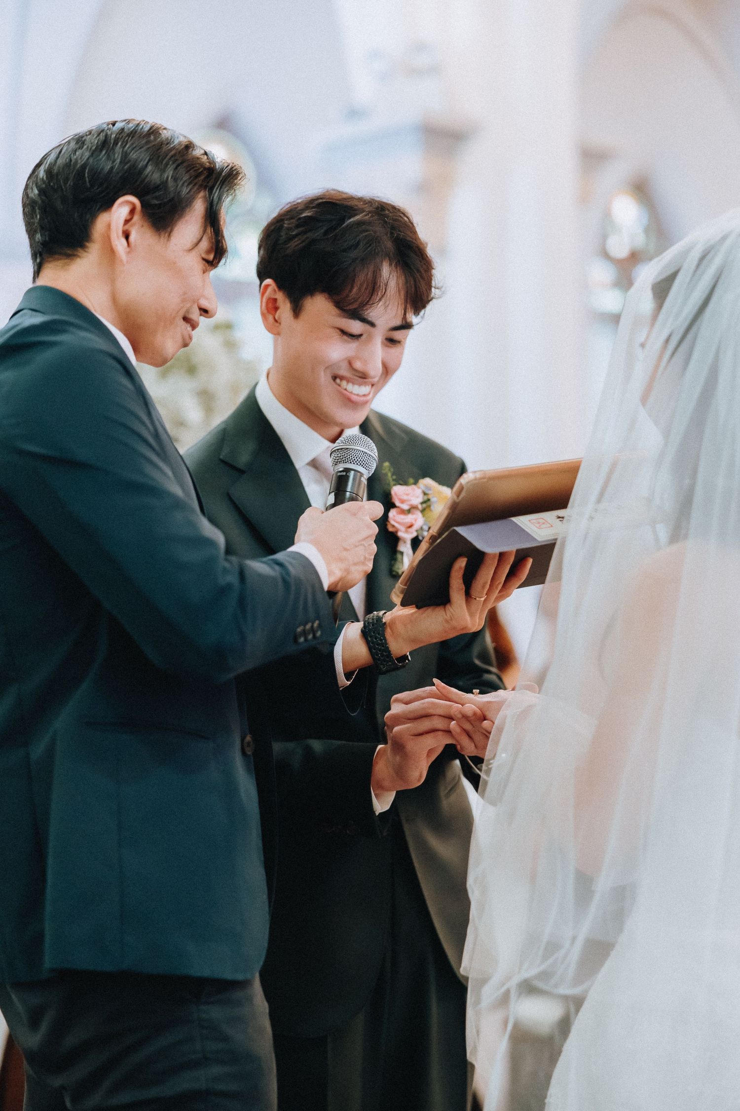 Smiling groom places wedding ring on bride's finger during church ceremony, assisted by best man holding microphone.