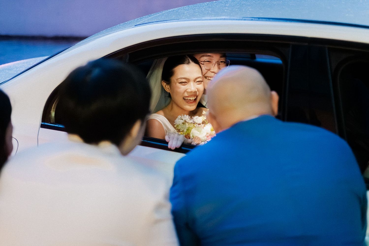 Smiling bride and groom inside white car greeted by guests during wedding day departure celebration.