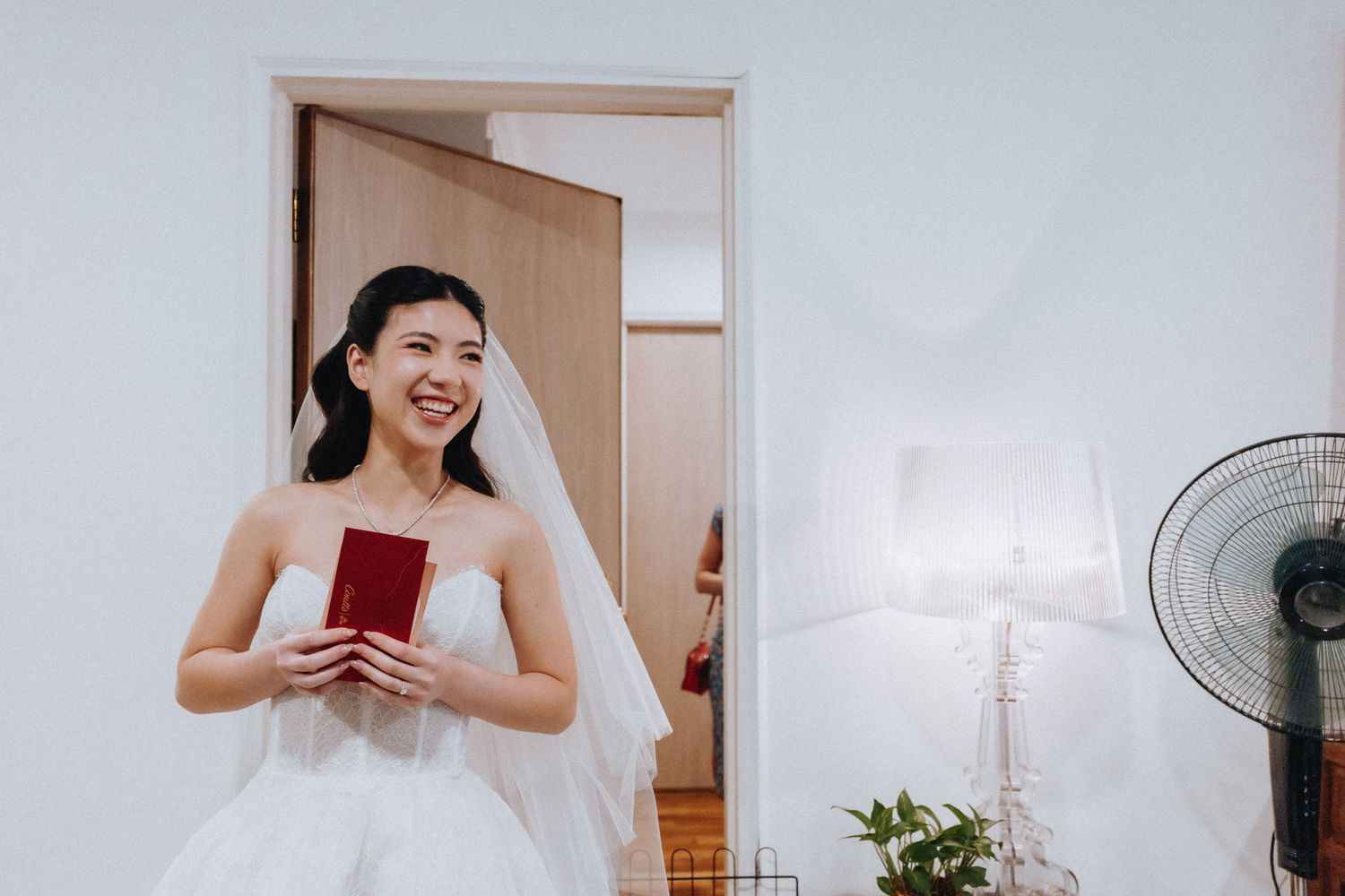 Smiling bride in white strapless gown and veil holding a red book, standing in a bright room with a fan nearby.