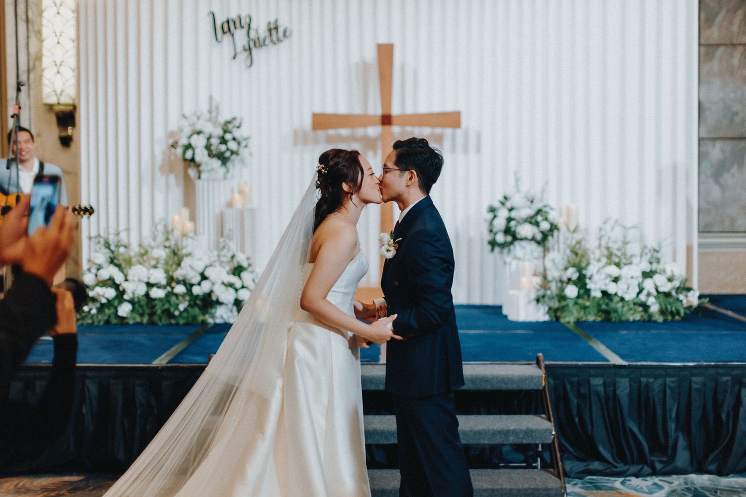 A wedding ceremony in front of a white wall with cross and floral arrangements.