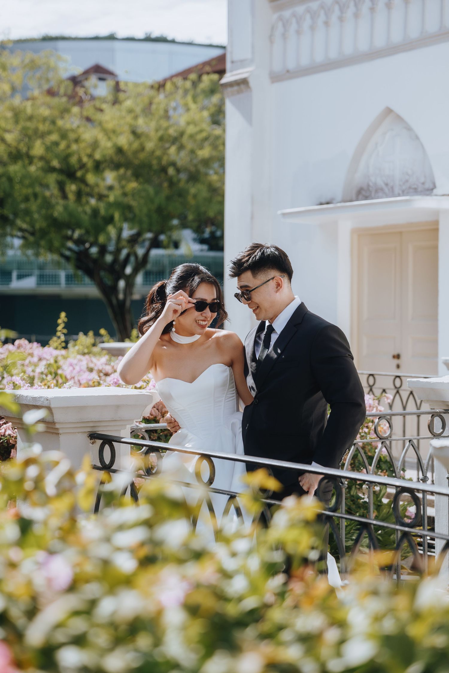 Bride and groom in sunglasses smiling together on a floral balcony outside a white chapel.