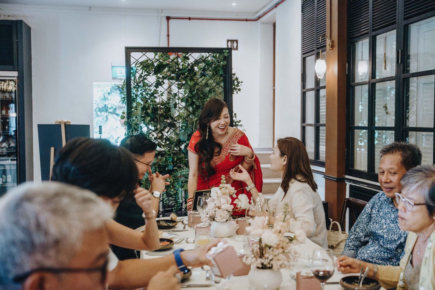 Woman in red dress smiles while greeting seated guests at a beautifully decorated wedding reception dinner.