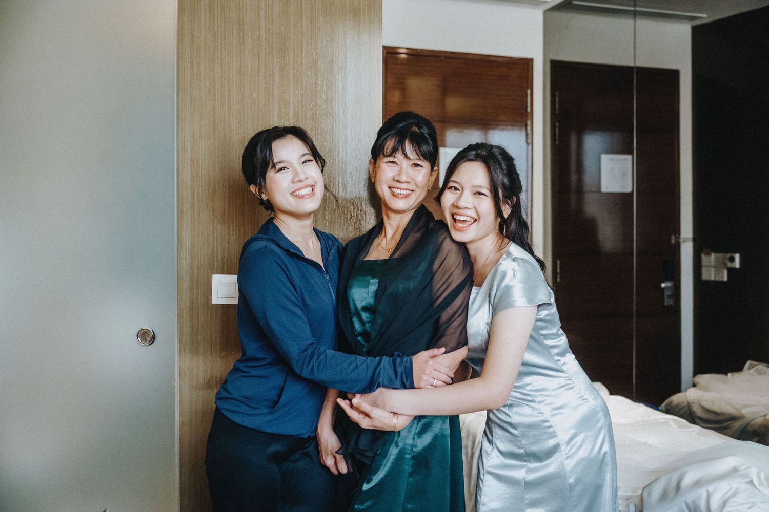 Three smiling Asian women in elegant dresses embrace warmly in a hotel room during wedding preparations.