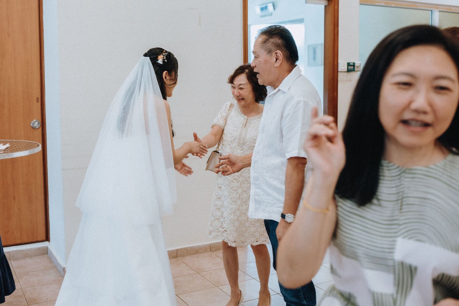 Asian bride in white wedding dress and veil greets elderly couple at indoor wedding celebration.