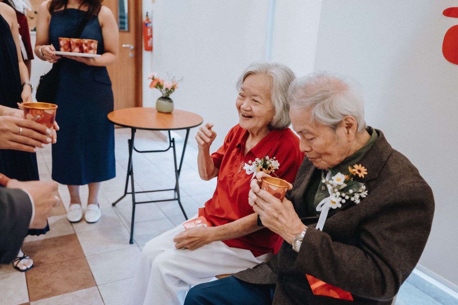 Two elderly women laughing joyfully at a wedding reception, holding floral corsages and festive drinks.