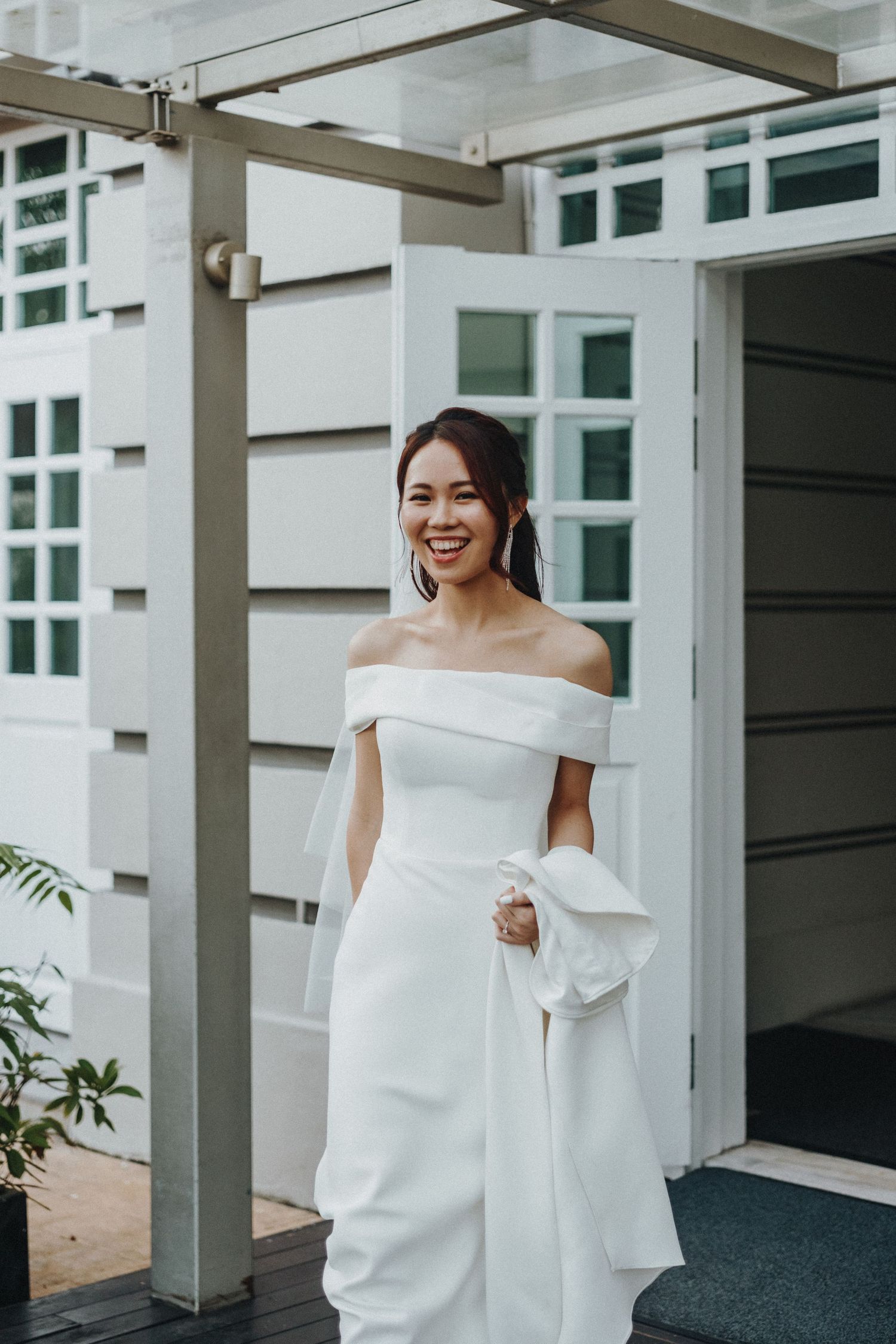 A bride wearing an elegant off-shoulder white wedding gown stands in front of a modern building entrance.
