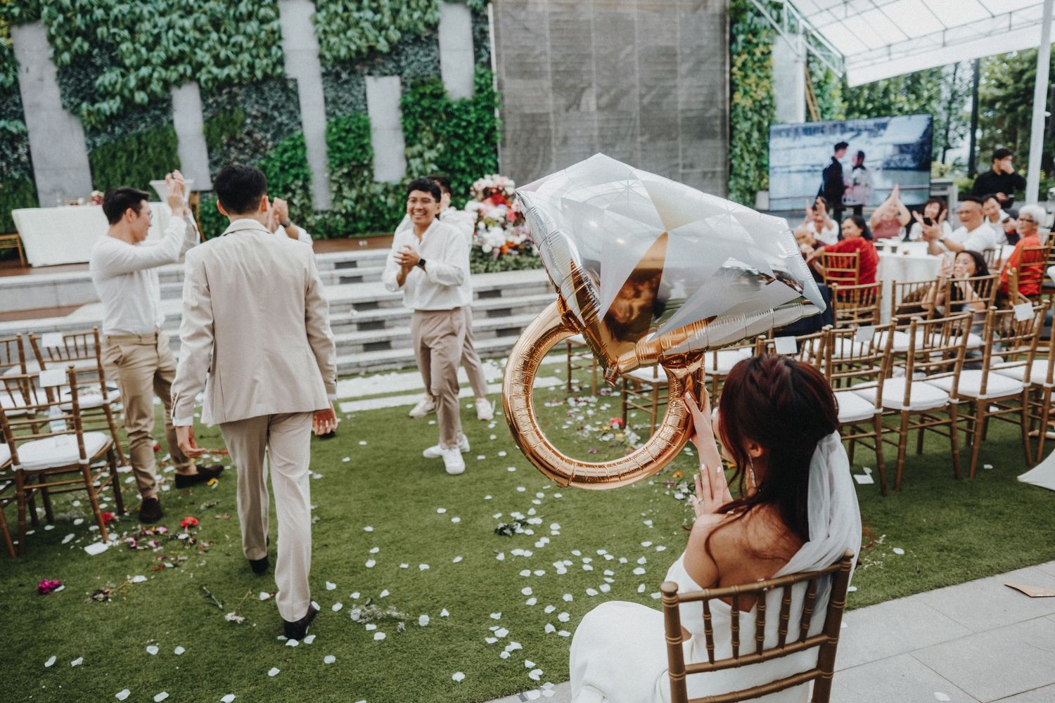 Celebratory scene at an outdoor wedding reception with rose petals scattered on the ground and wooden chairs lined up.