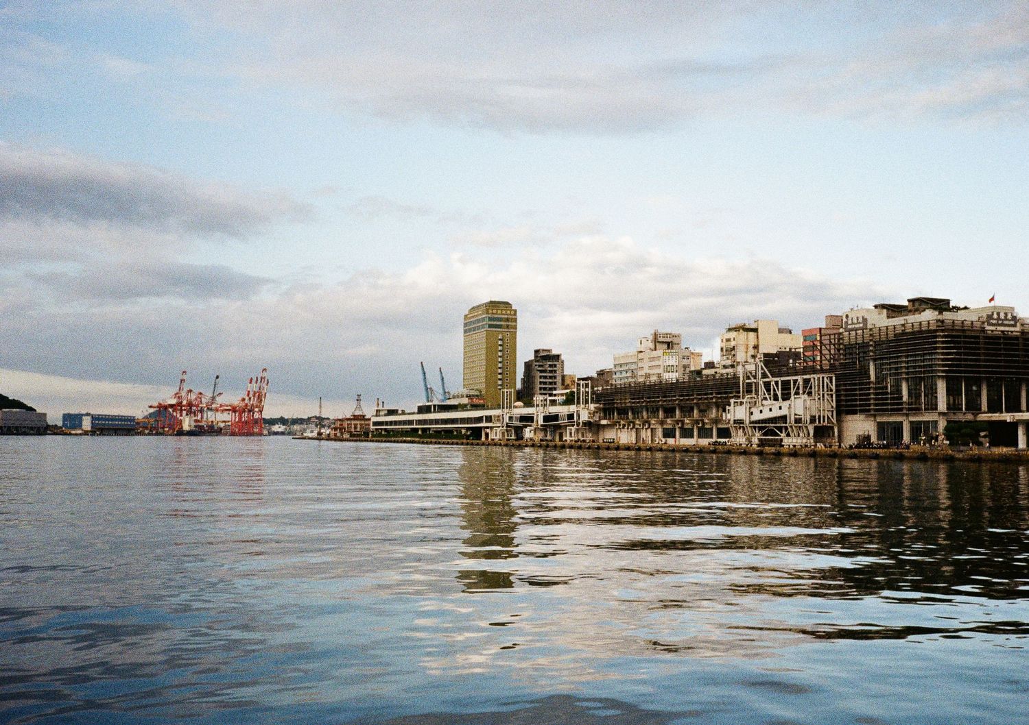 Waterfront cityscape with buildings and shipping docks reflected in calm harbor waters.