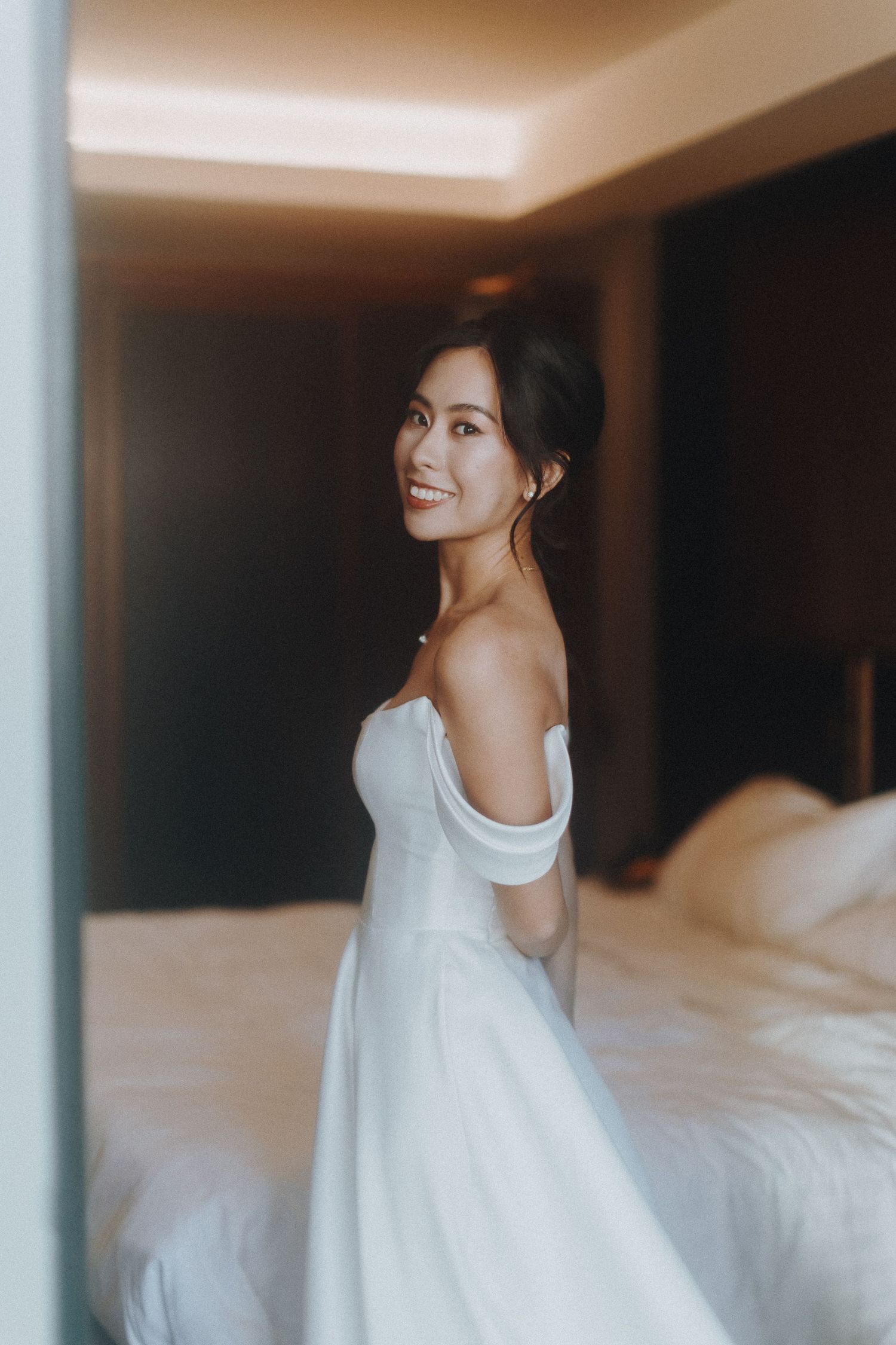A bride in an elegant off-shoulder white wedding dress poses in a softly lit hotel room.