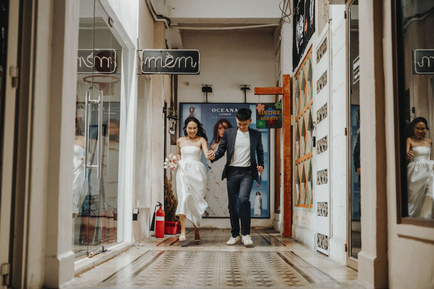 A couple in wedding attire walks hand in hand down a bright shopping arcade lined with mirrors and storefronts.