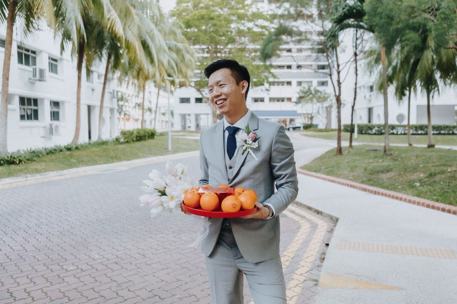 Wedding attendant in grey suit holding traditional mandarin oranges on a palm-lined pathway.