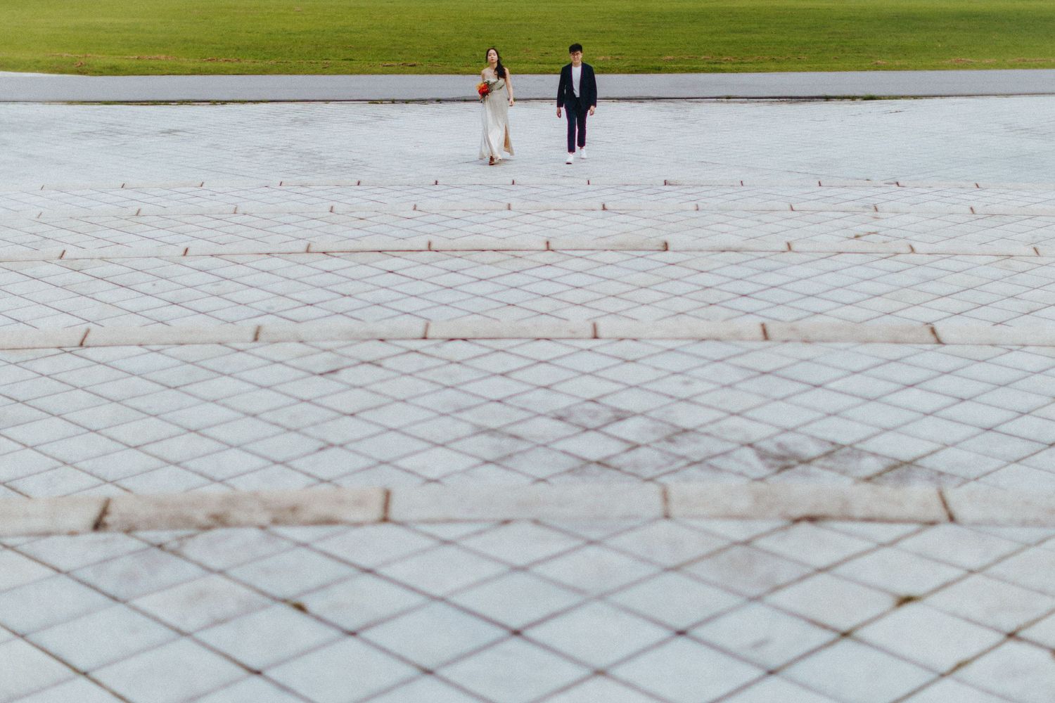 A couple stands together on a geometric patterned stone plaza with green grass in the background.