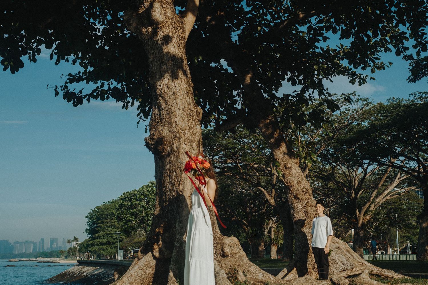 Couple poses creatively around a large tree trunk near the beach with a red floral bouquet.