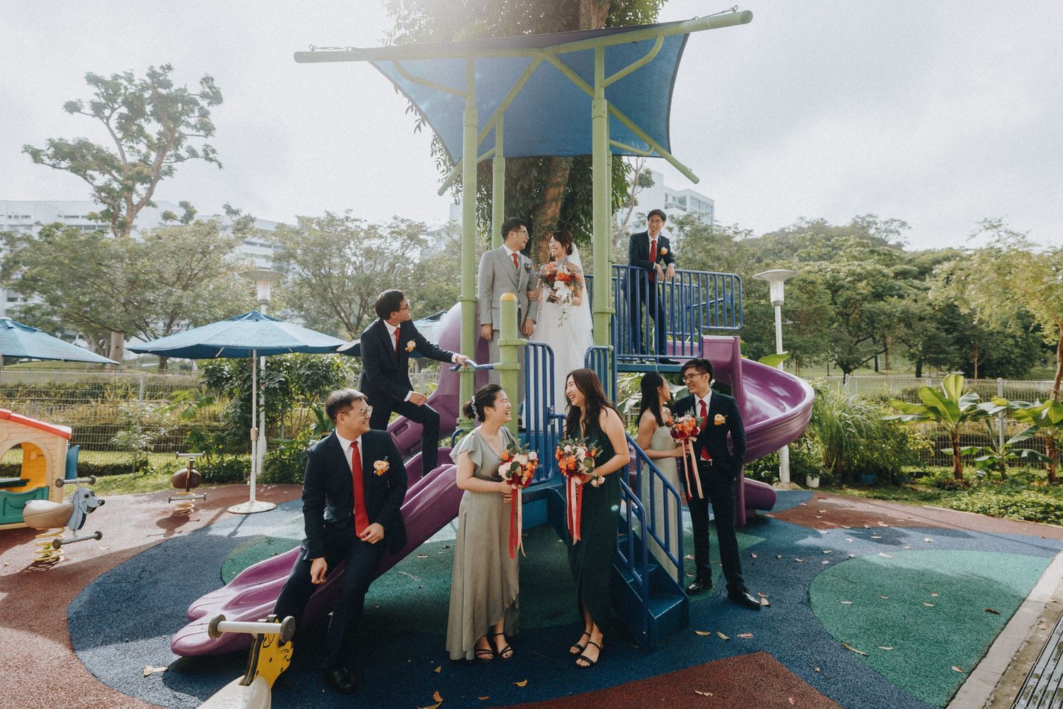 Wedding party poses playfully on a colorful playground structure outdoors.
