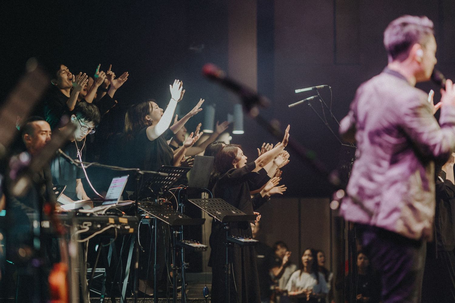 Musicians perform on stage with raised hands visible in the background during a worship service.