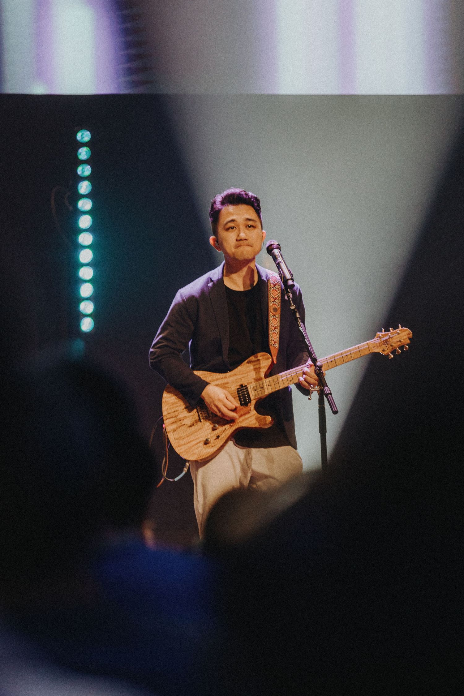 A performer plays an electric guitar on stage with blue accent lighting and vertical lights in the background.