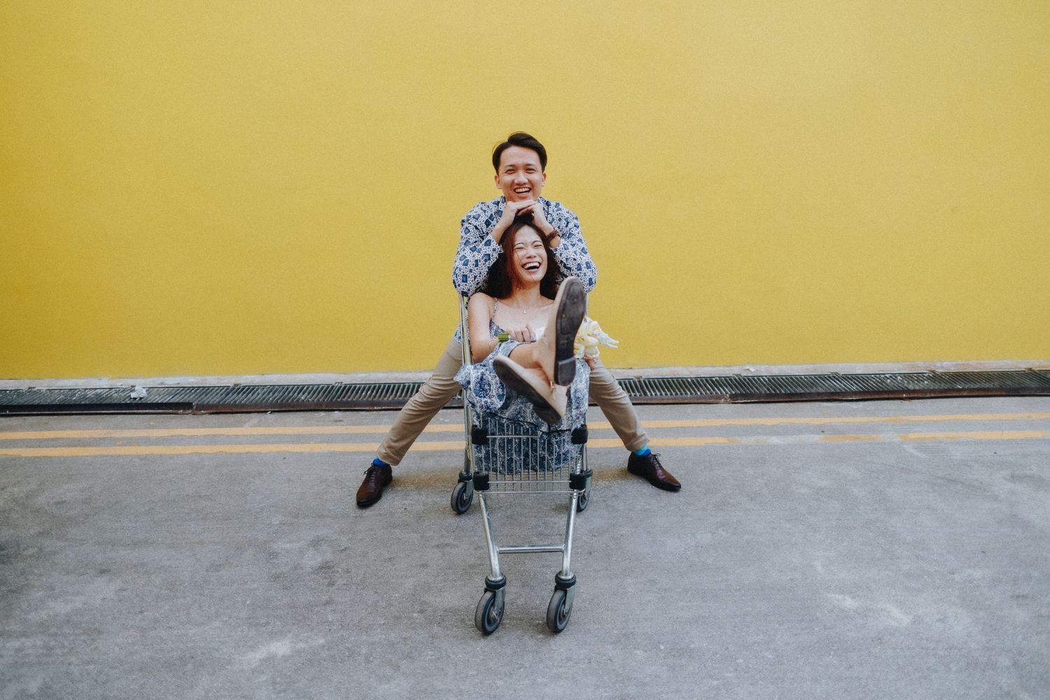 Playful couple poses with shopping carts against a bright yellow wall in matching outfits.