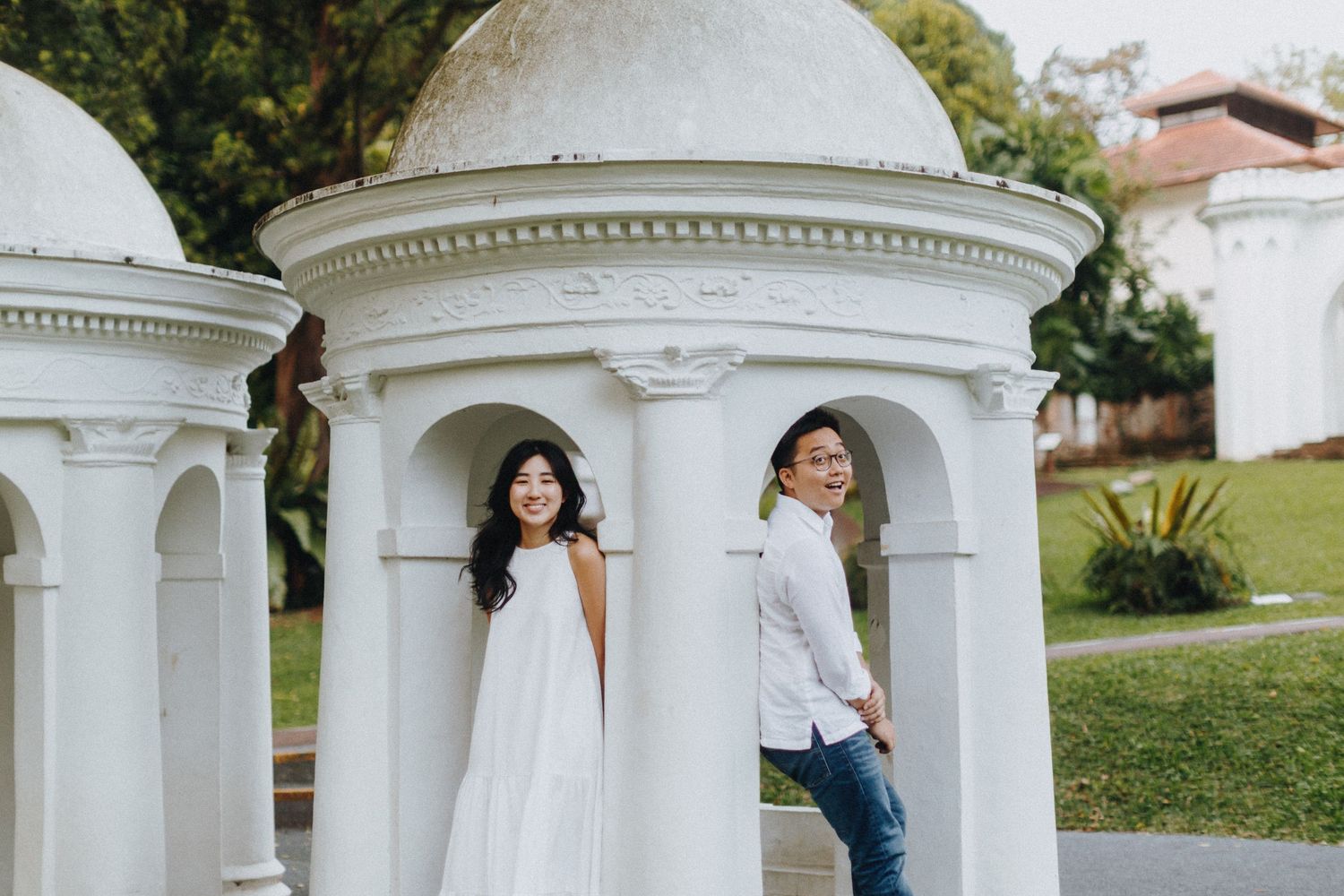 A couple poses playfully between white classical architecture columns in a garden setting.