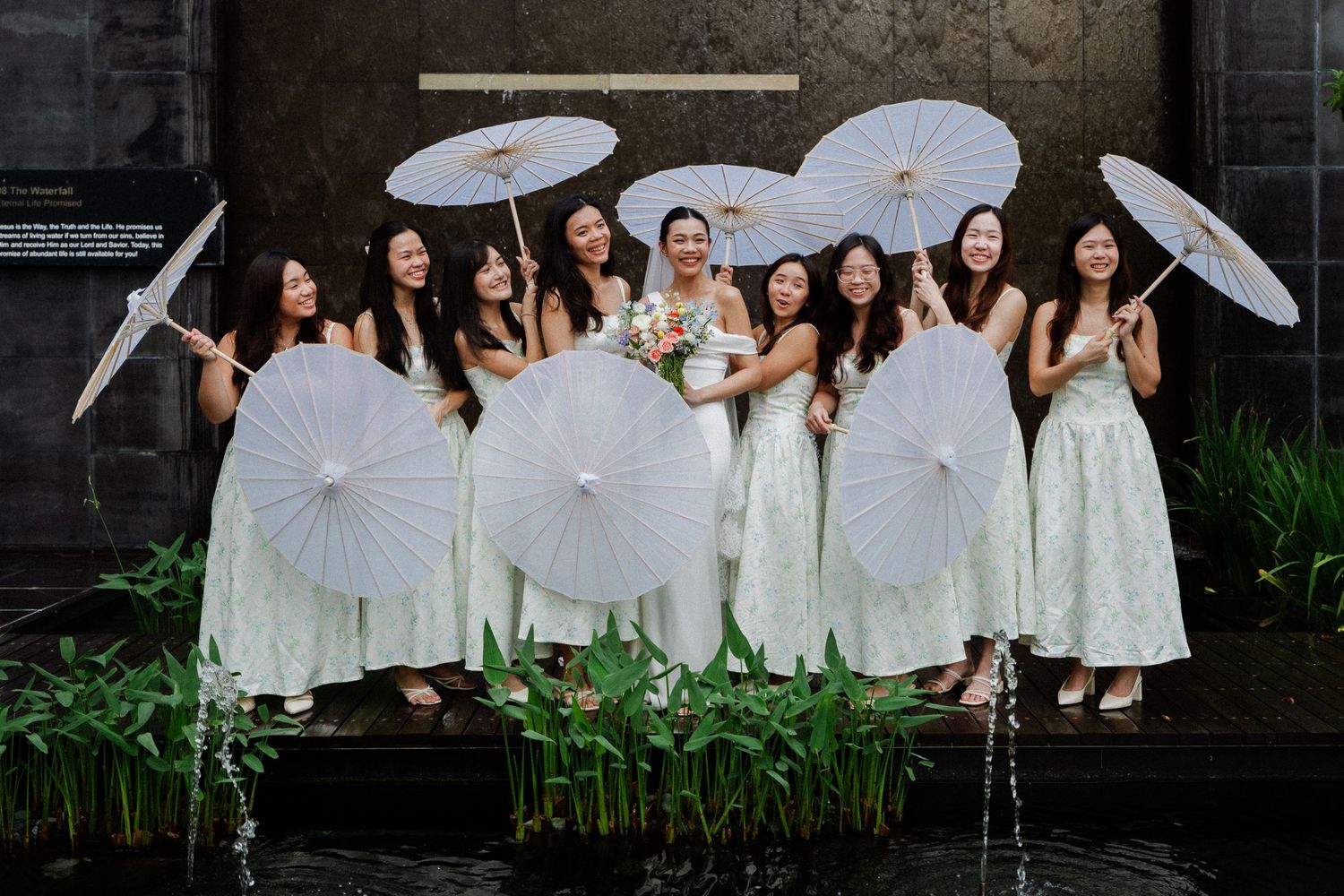 Bride and bridesmaids in light green dresses holding white parasols outdoors near water feature.