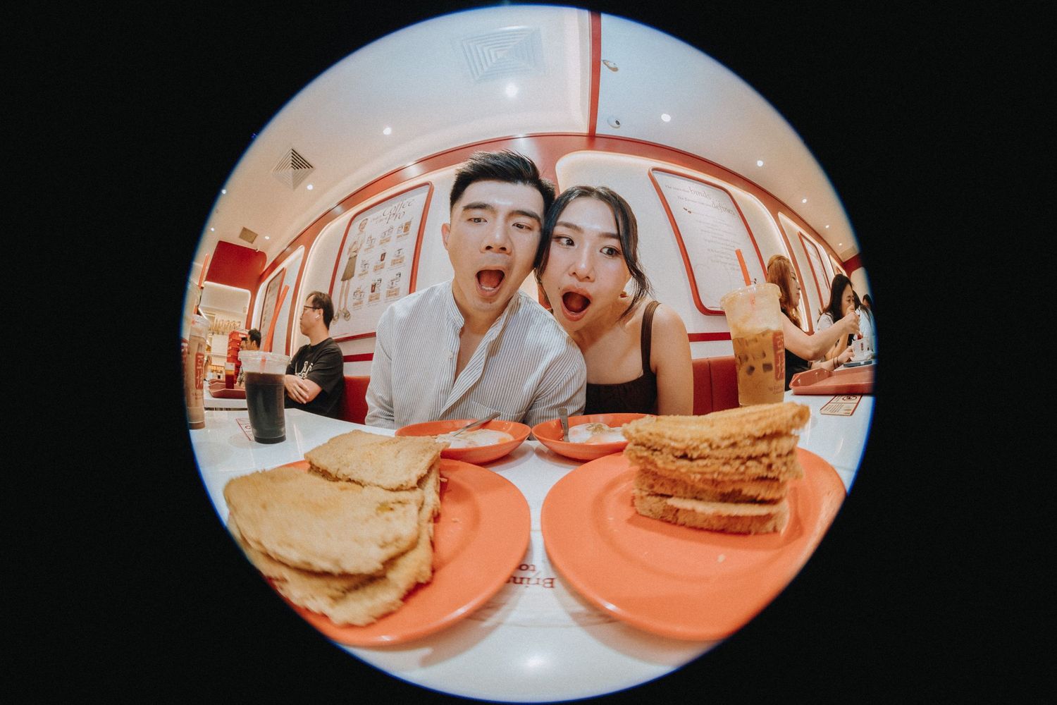 Couple making funny faces at a restaurant table with toast and sandwiches on orange plates, shot with fisheye lens.