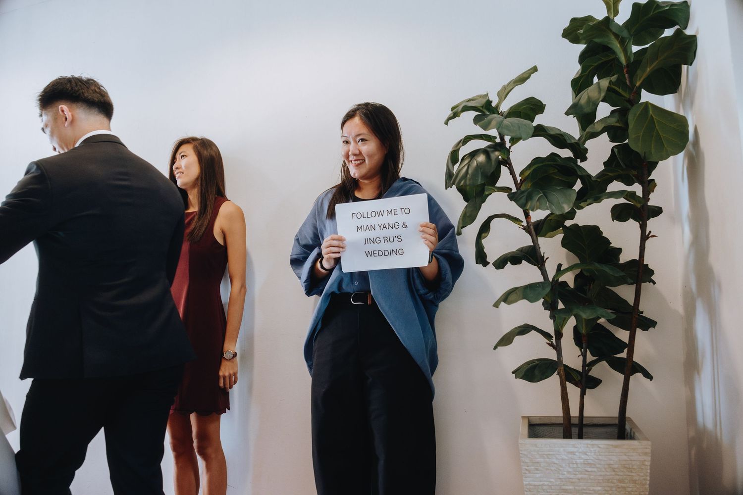 Woman holds a sign directing guests to a wedding ceremony while attendees gather in a bright modern venue.