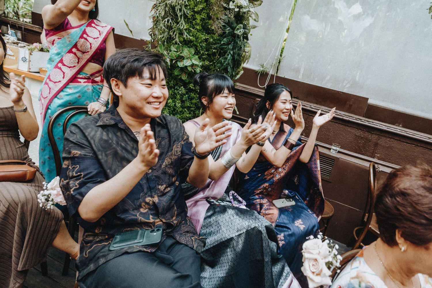 Joyful wedding guests in traditional attire clapping and celebrating during an outdoor garden wedding reception.