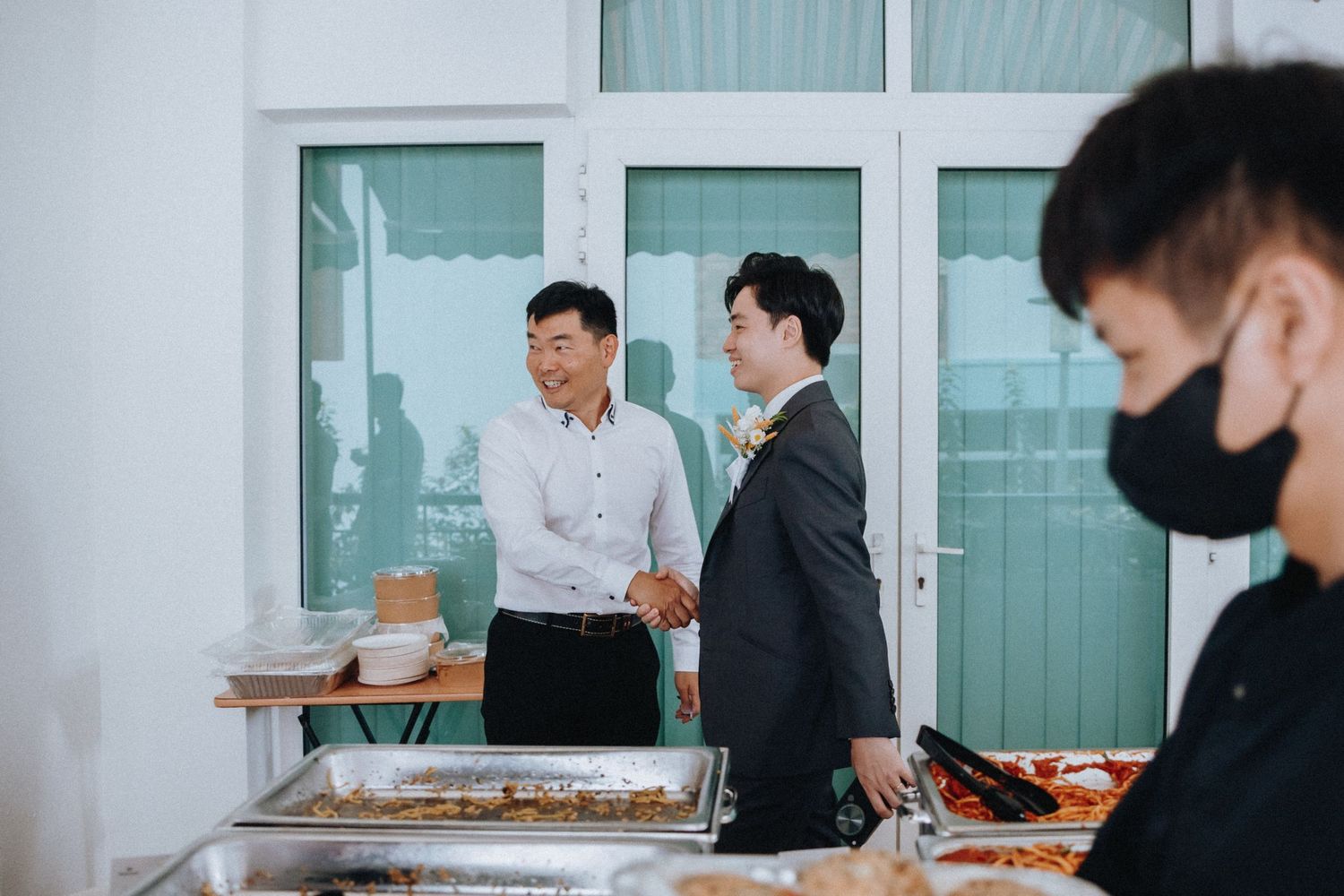 Two men shake hands warmly near a buffet table at a wedding reception celebration.