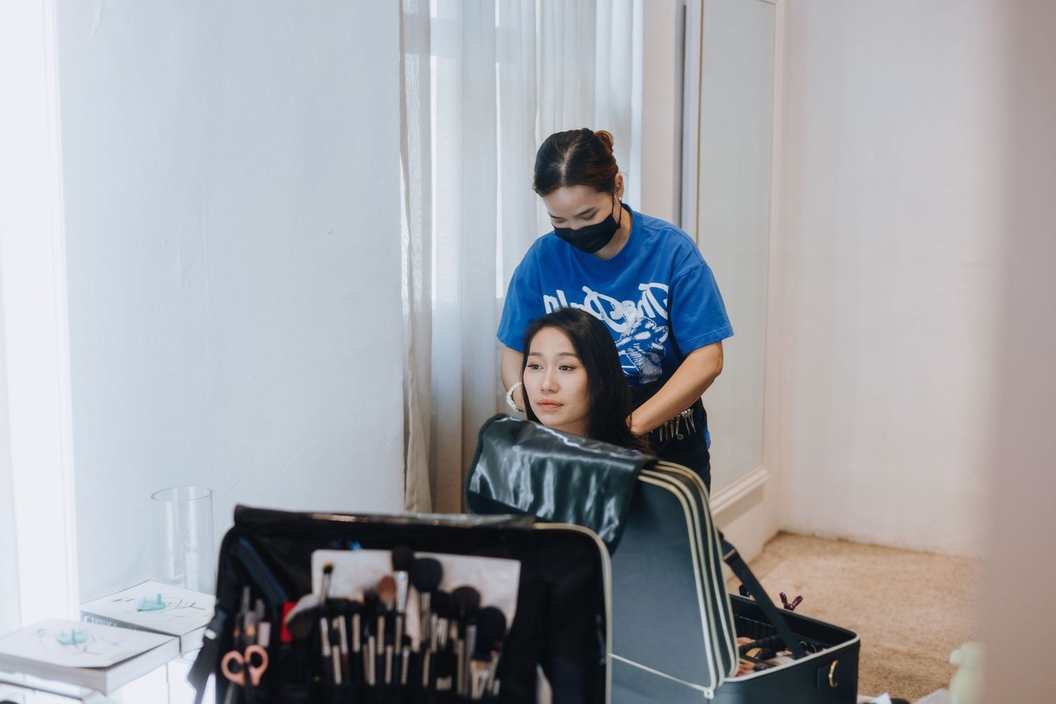 Makeup artist in blue shirt and mask styling a client's hair in a bright salon studio with brushes on display.