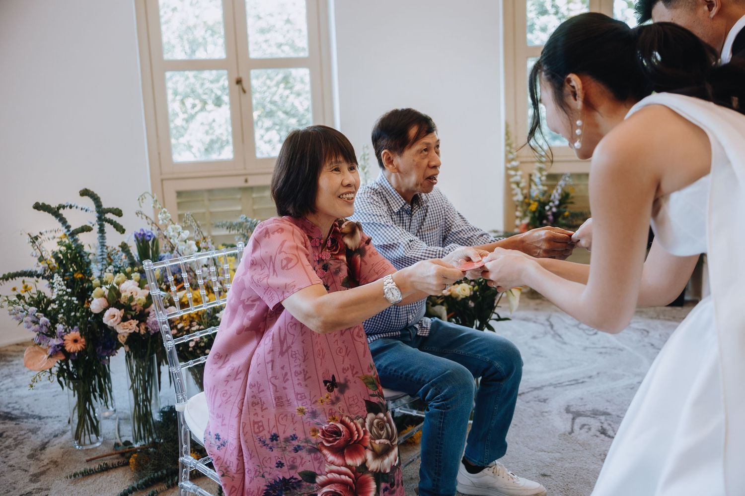 Bride in white gown receives gift from smiling guest seated near floral wedding decorations.