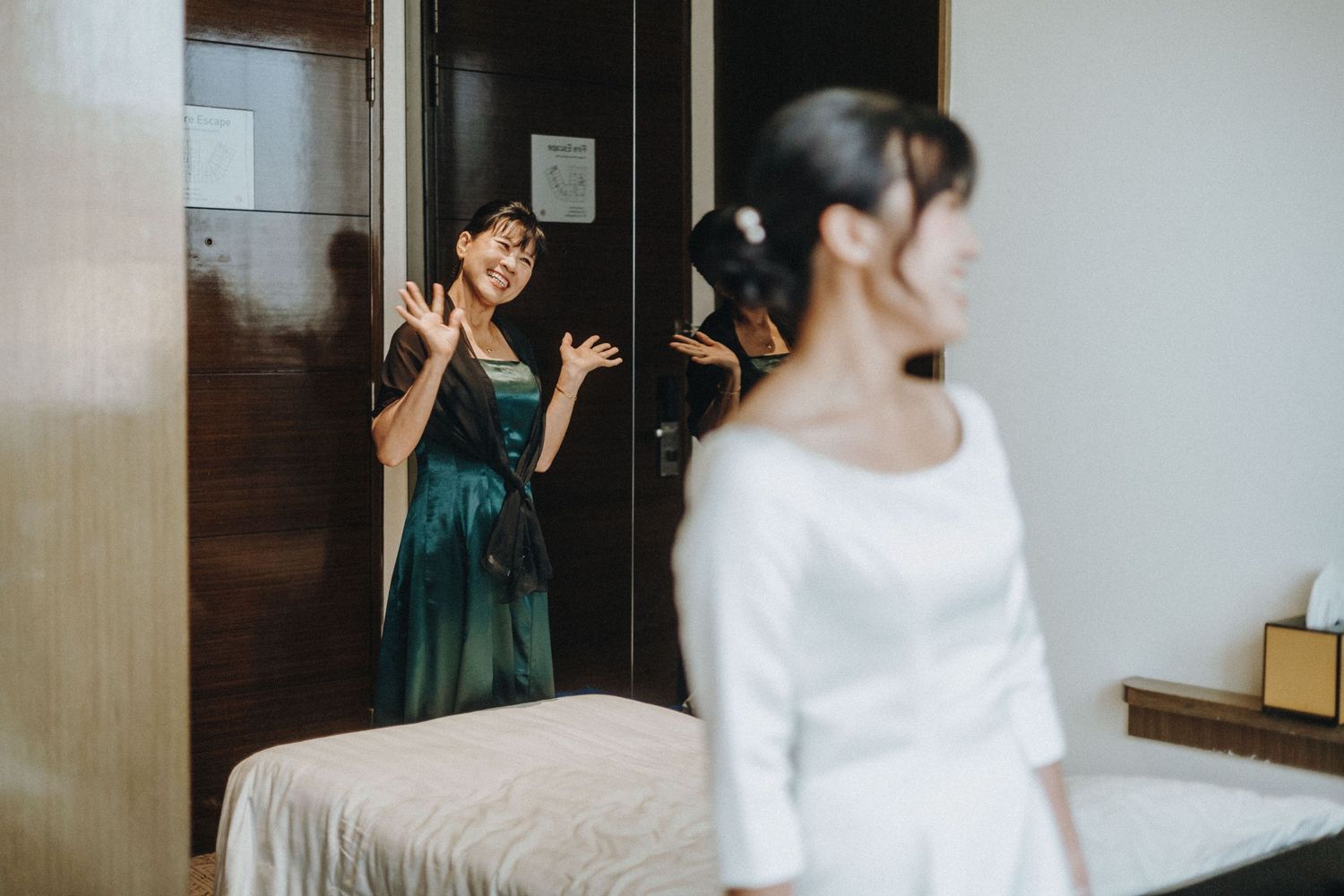 A woman in a green dress reacts with joy upon seeing a bride in a white gown during wedding preparations indoors.