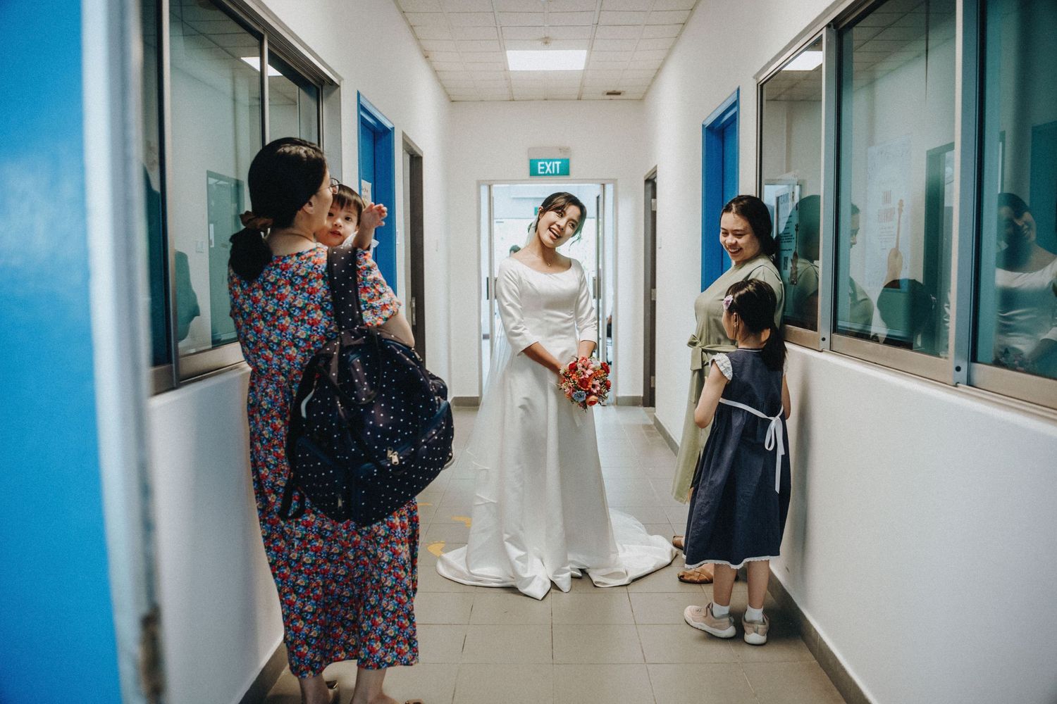 Bride in white dress greeted by family and child in bright hospital corridor hallway.