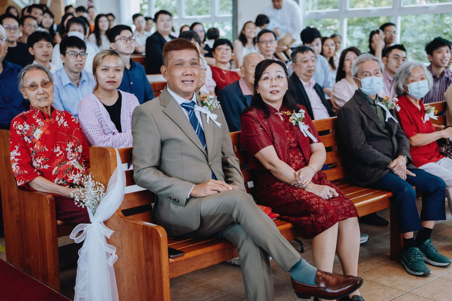 A groom in a tan suit sits with family members in church pews during a wedding ceremony.