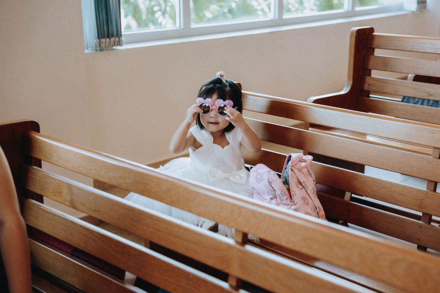 Adorable little girl in white dress playing with pink toys on church pew.