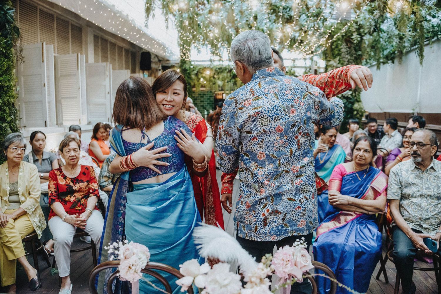 Wedding guests in vibrant saris share warm embraces during an outdoor multicultural ceremony reception.