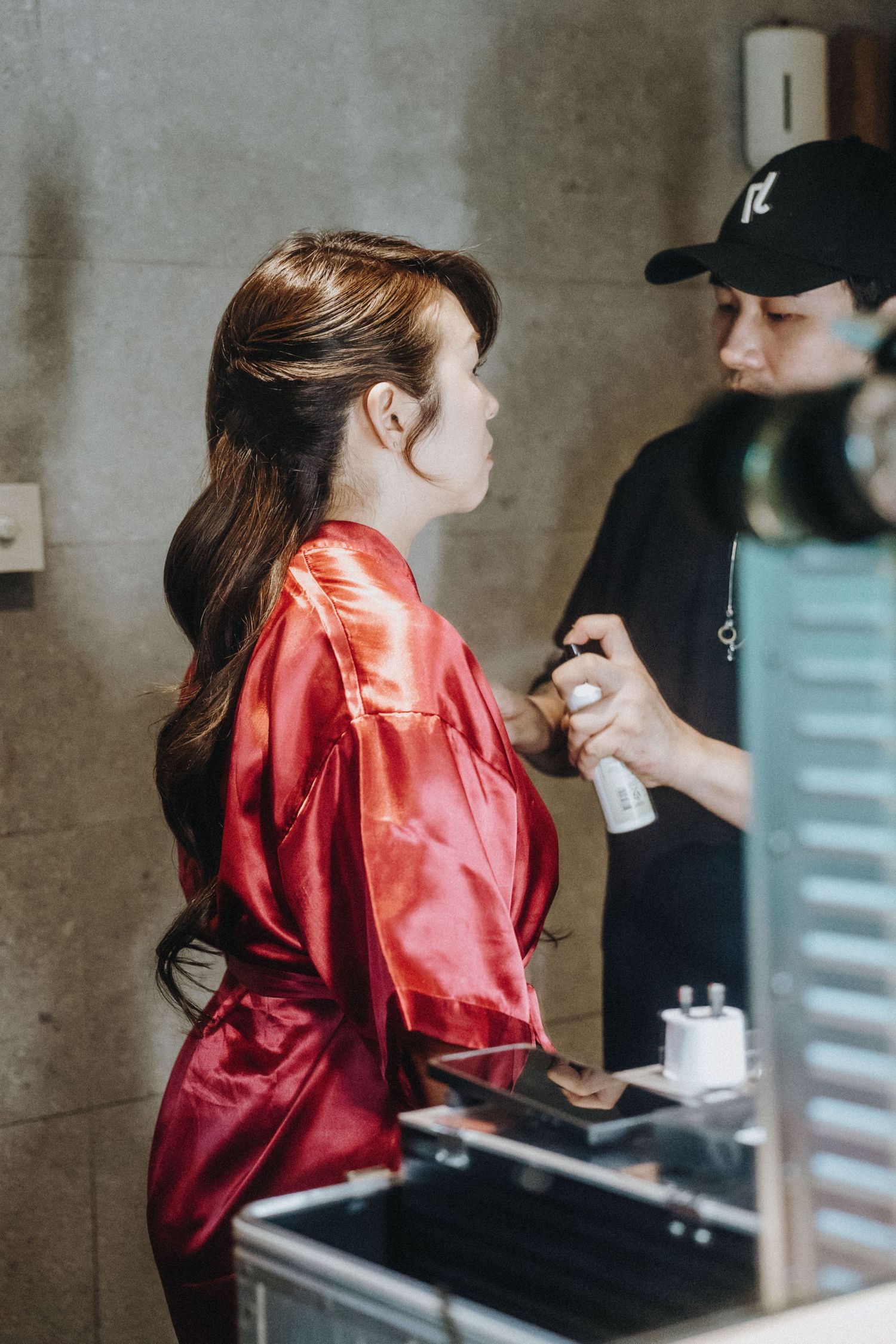 A hairstylist applying hair spray to a woman in a red satin robe during bridal preparation.