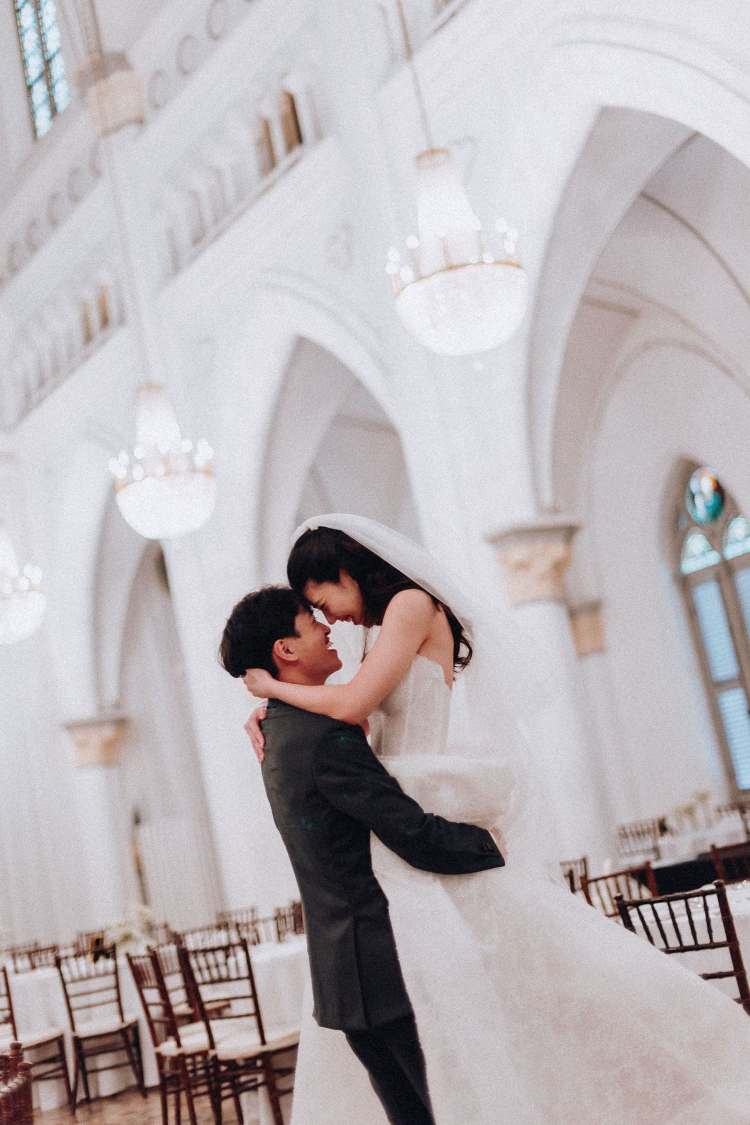Groom lifting bride in white gown for a romantic moment inside grand white cathedral with chandeliers.
