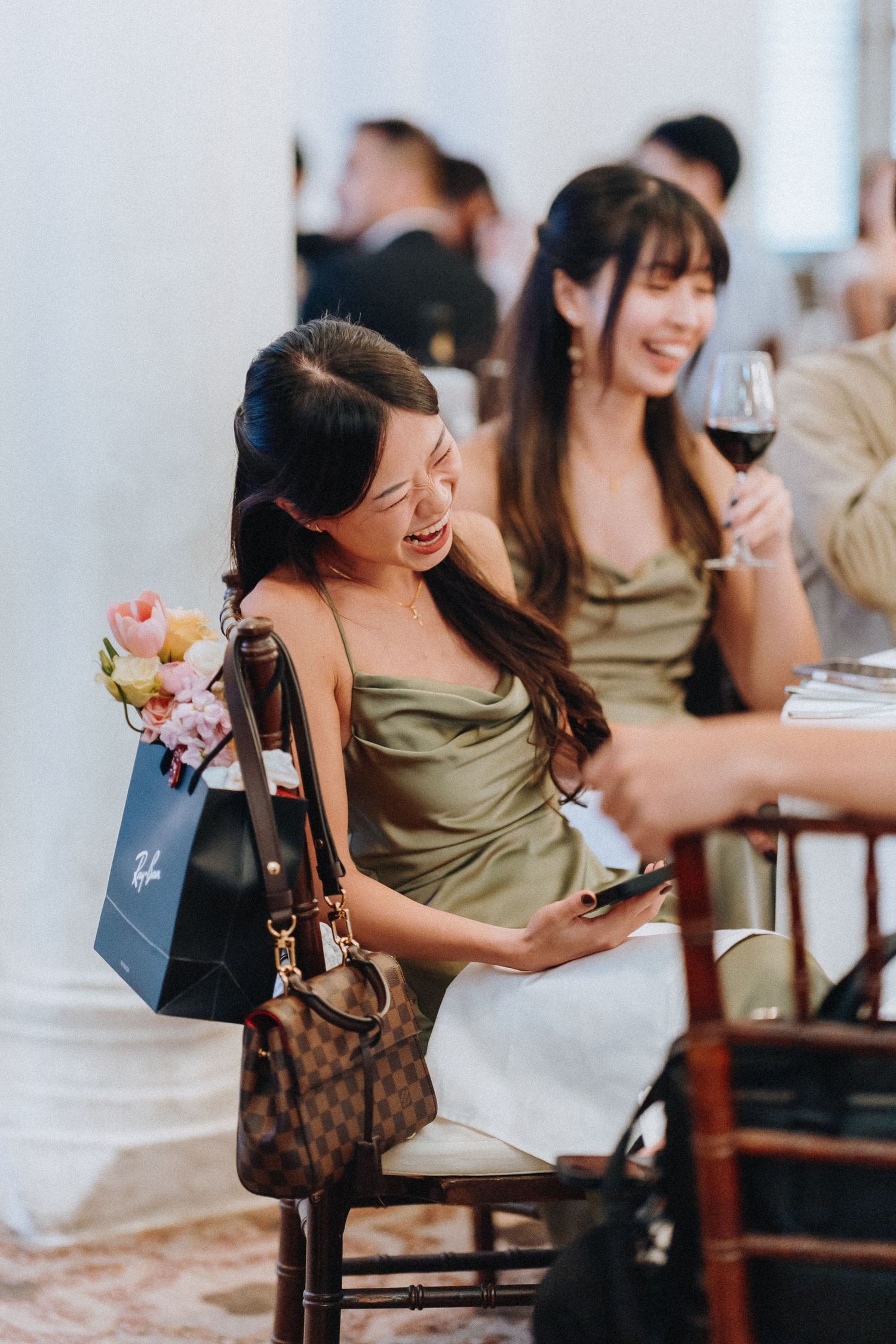 Woman in olive dress laughing at a wedding reception while holding a phone and Louis Vuitton bag.