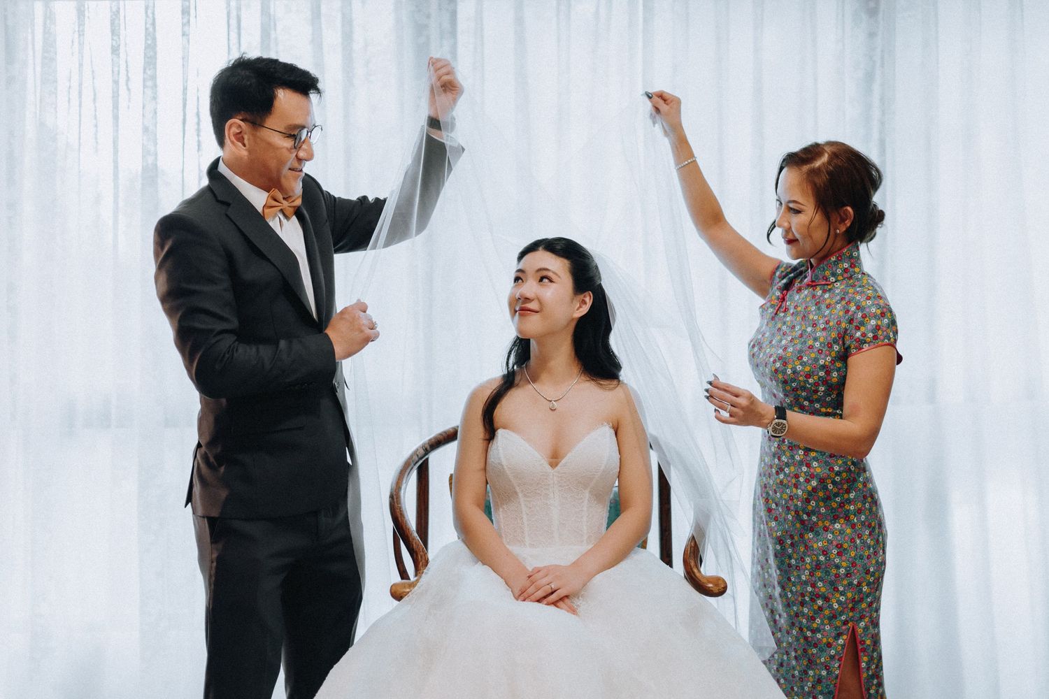 Parents helping bride with veil in white wedding dress during bridal preparation ceremony.