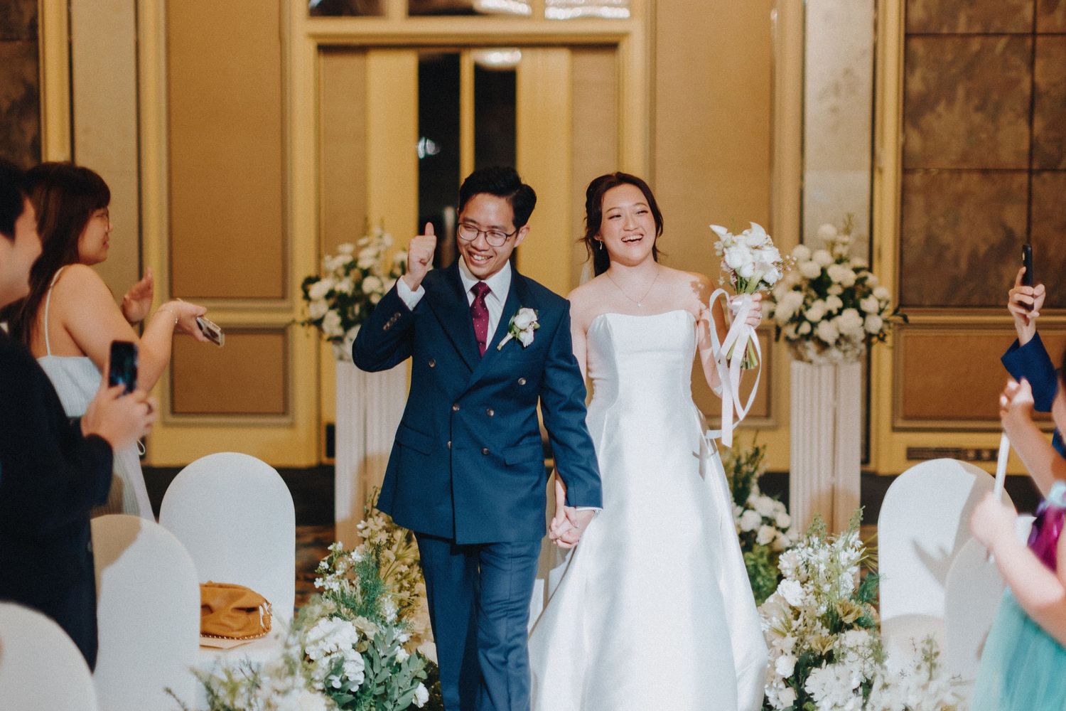 A joyful celebration as newlyweds walk down the aisle decorated with white flowers and chair covers at their wedding ceremony.