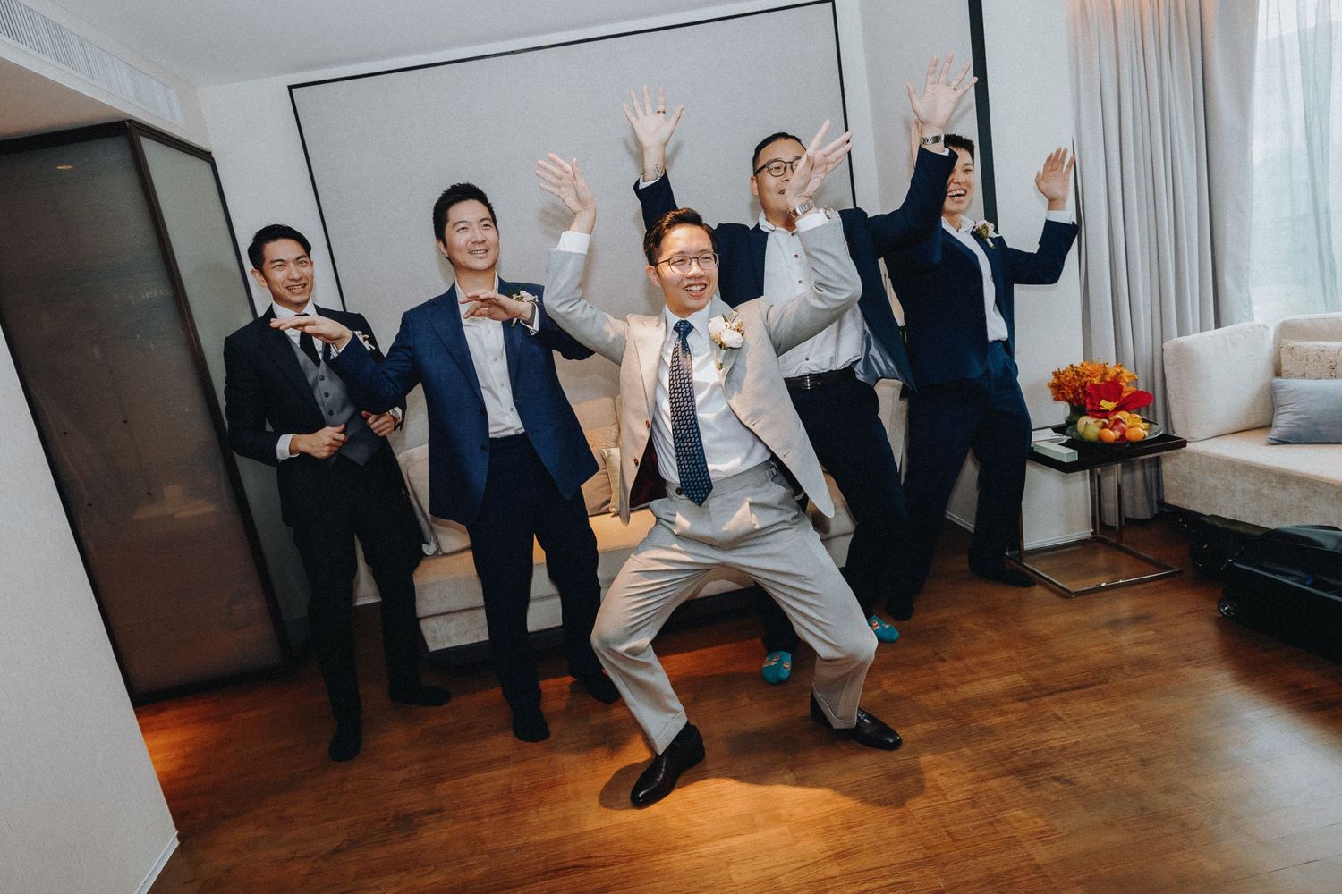 Wedding party members jumping and celebrating in formal attire in an indoor venue.