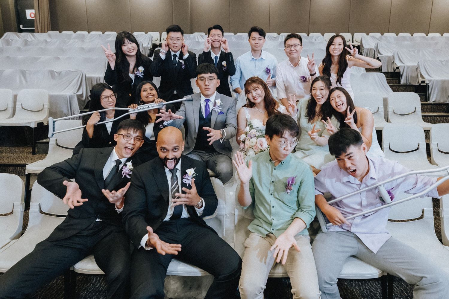 Wedding party members laugh together while sitting on white chairs during outdoor ceremony.