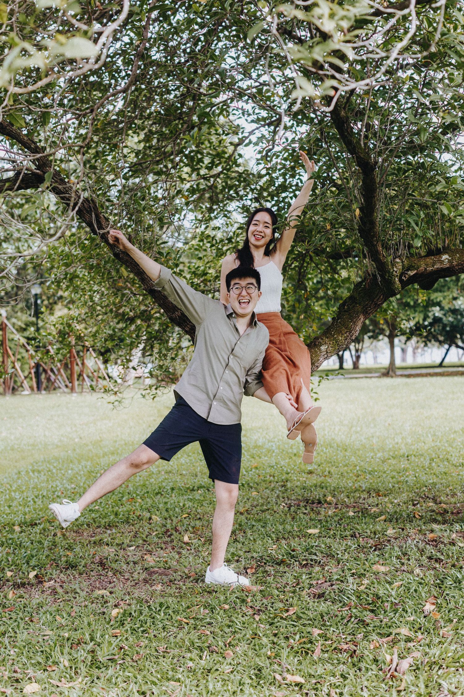 Dancing figures leap joyfully beneath tree branches in an outdoor park setting on a bright sunny day.