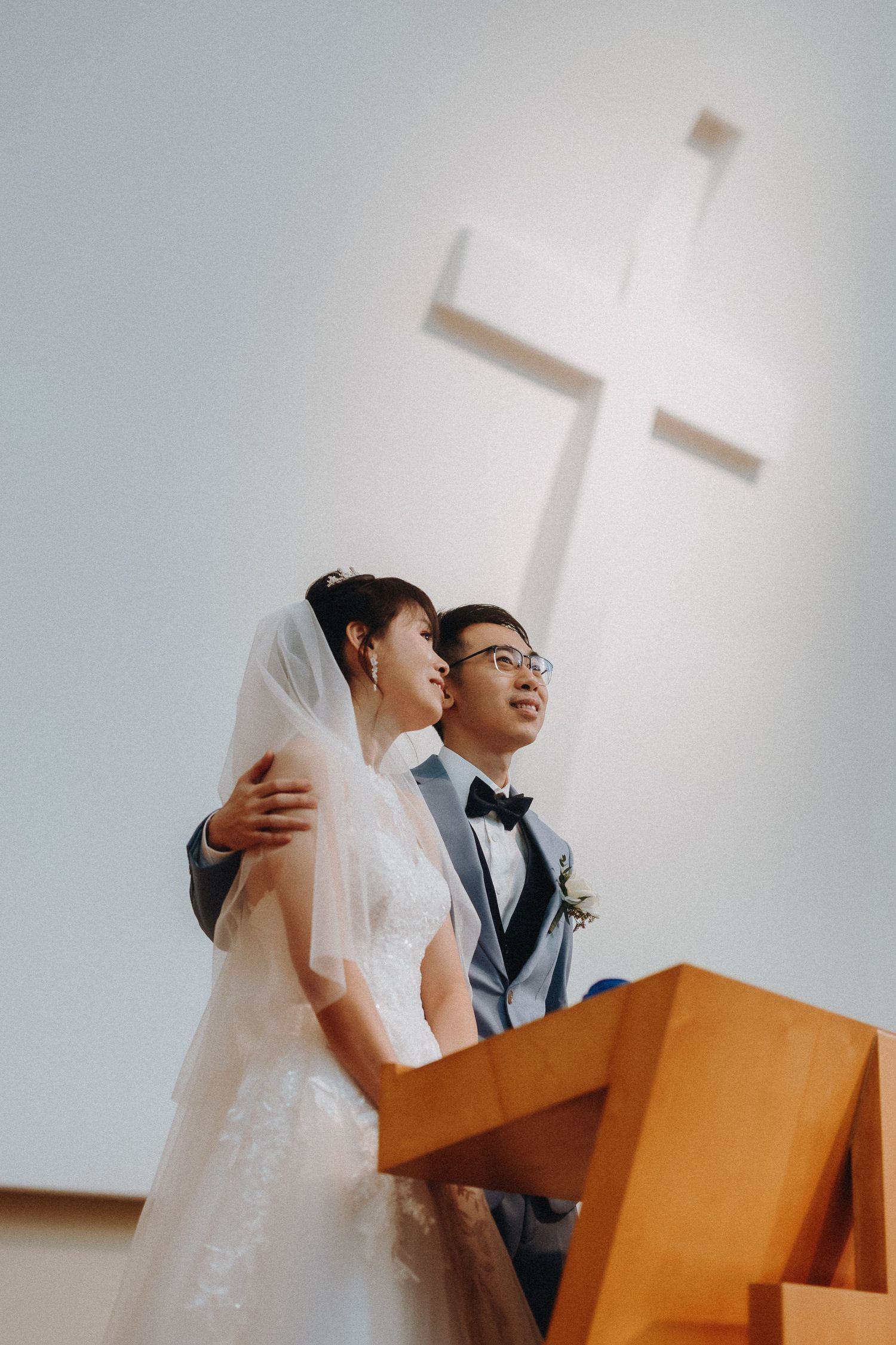A couple stands together at a church altar with a wooden podium and white cross visible in the background.